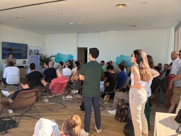 An indoor scene showing an audience gathered for a presentation, with a speaker standing near a screen on the left.