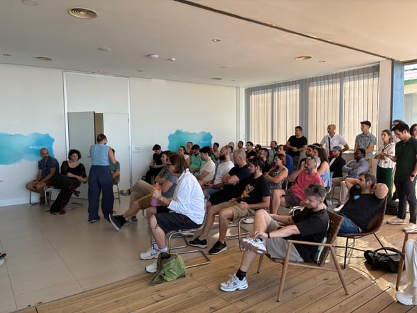 A group of people are seated in an indoor space, listening to a presentation delivered by a speaker standing near the front wall.