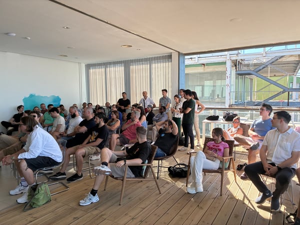 A group of people are gathered in a modern, open-sided room with wooden flooring, attentively watching a presentation.