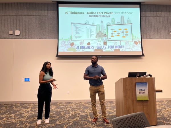 A photograph capturing an indoor professional event where a woman and a man are presenting in front of a large projection screen displaying a title slide for the 'AI Tinkerers - Dallas Fort Worth' meetup.
