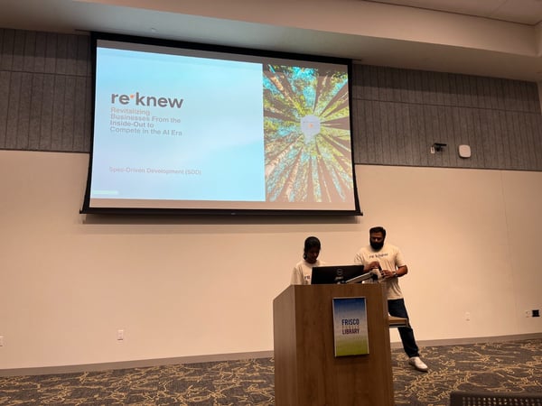 A photograph capturing an indoor presentation where two individuals are standing near a wooden podium in front of a large projection screen displaying a title slide for a talk about revitalizing businesses in the AI era.