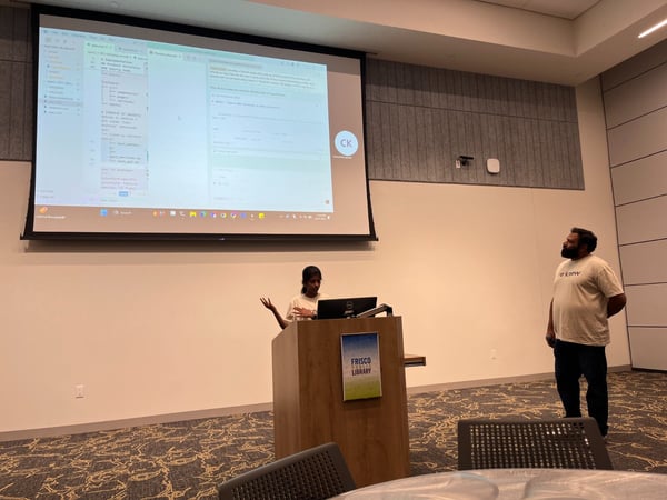 An indoor photograph capturing a technical presentation where a woman speaks from a wooden lectern labeled 'FRISCO LIBRARY,' while a large screen displays computer code and documentation to the audience, and a man stands observing the screen on the right.