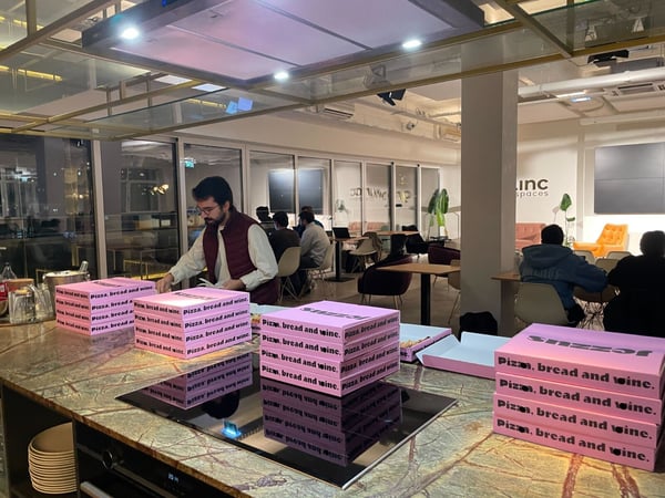 A man wearing a maroon vest stands behind a marble kitchen island stacking bright pink pizza boxes labeled "Pizza. bread and wine." while several people are seated at tables in the background of a modern co-working space.