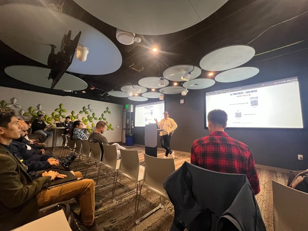 A photograph taken indoors shows a technology or business presentation event, where a speaker stands near a podium addressing an audience seated in rows. The room features modern decor, including circular ceiling panels and green acoustic wall panels, with a large screen displaying the title "AI TINKERERS - SAN DIEGO."