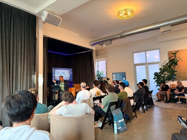 An indoor presentation event where a speaker stands next to a screen displaying slides, addressing an audience seated in chairs.