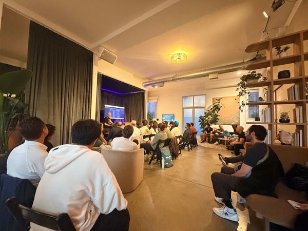 A photograph capturing an indoor presentation or meetup event, with an audience seated in a warmly lit, modern room looking towards a speaker and screen on a small stage.
