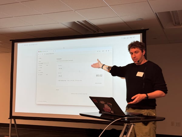 A man stands next to a large projector screen, gesturing towards a currency conversion web application displayed on it during a presentation.