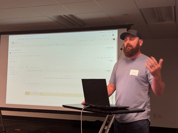 A man with a beard and a baseball cap is giving a presentation, gesturing with his hand next to a laptop on a stand with a large projector screen displaying a GitHub commit in the background.