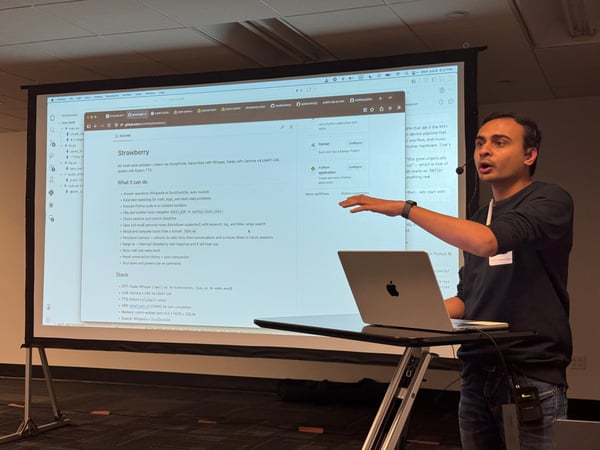A man stands next to a laptop on a stand, gesturing with his hand as he presents a project called 'Strawberry' on a large projection screen behind him.