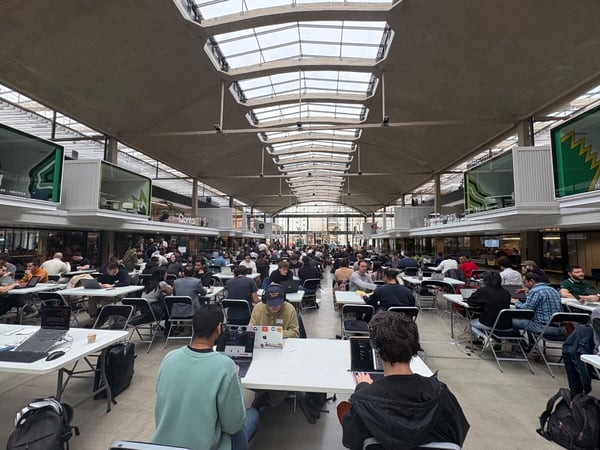 A wide-angle photograph capturing a large indoor event, likely a hackathon or co-working session, where dozens of people are seated at long rows of white tables working on laptops under a high ceiling with skylights.