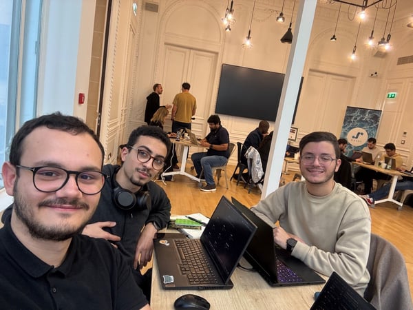 A selfie taken by three young men working on laptops in the foreground, while several other people are visible working or talking in the background of a large, high-ceilinged room.
