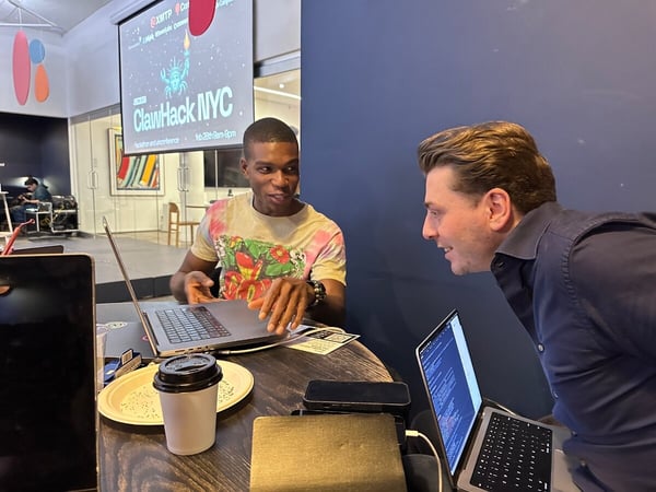 Two men sit at a table with laptops, engaged in a conversation. A projection screen in the background displays 'ClawHack NYC' branding for a hackathon event.