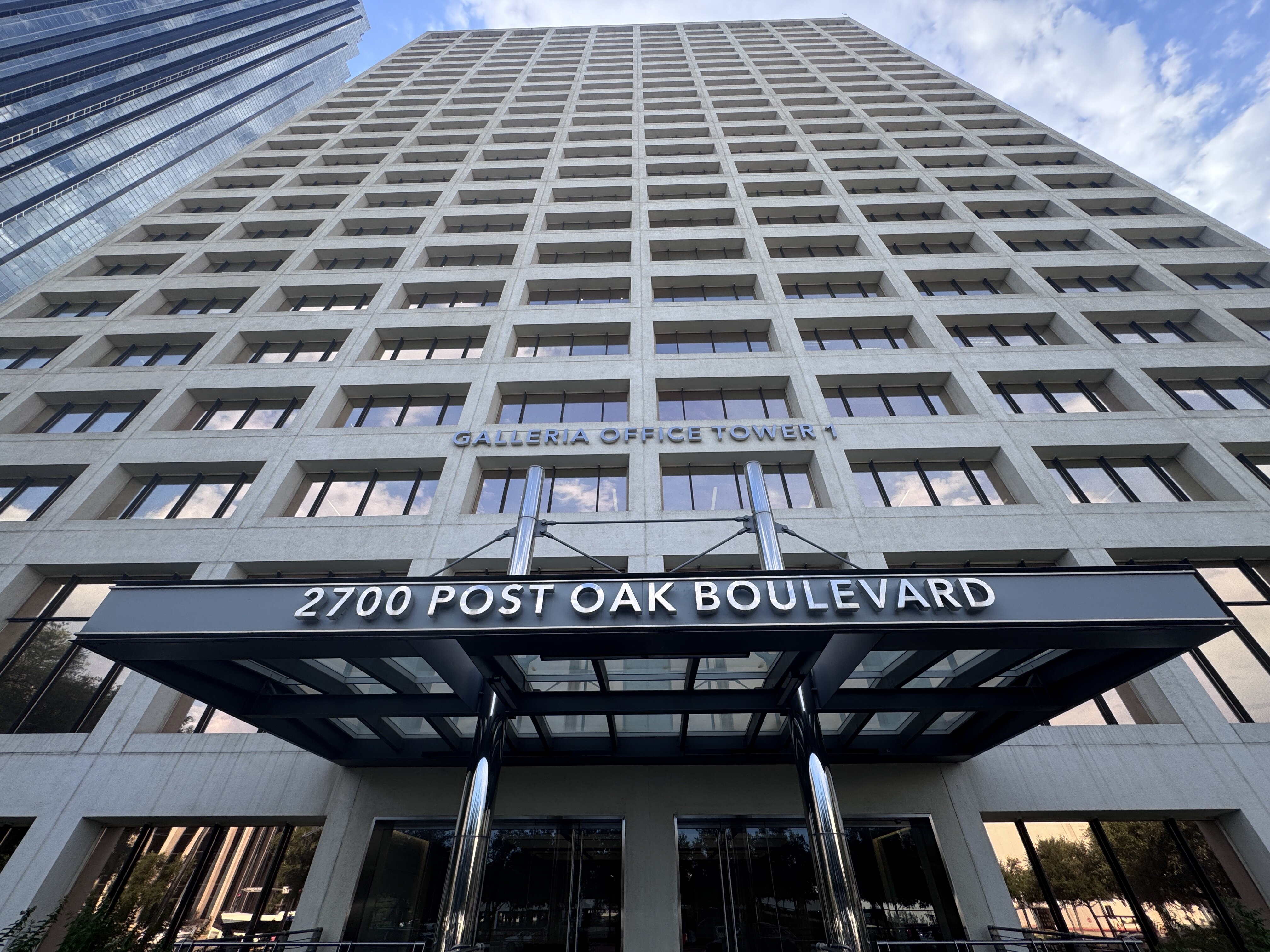 A low-angle photograph captures the imposing facade of a tall, grid-patterned office building, identified by signage as "Galleria Office Tower 1" located at "2700 Post Oak Boulevard."