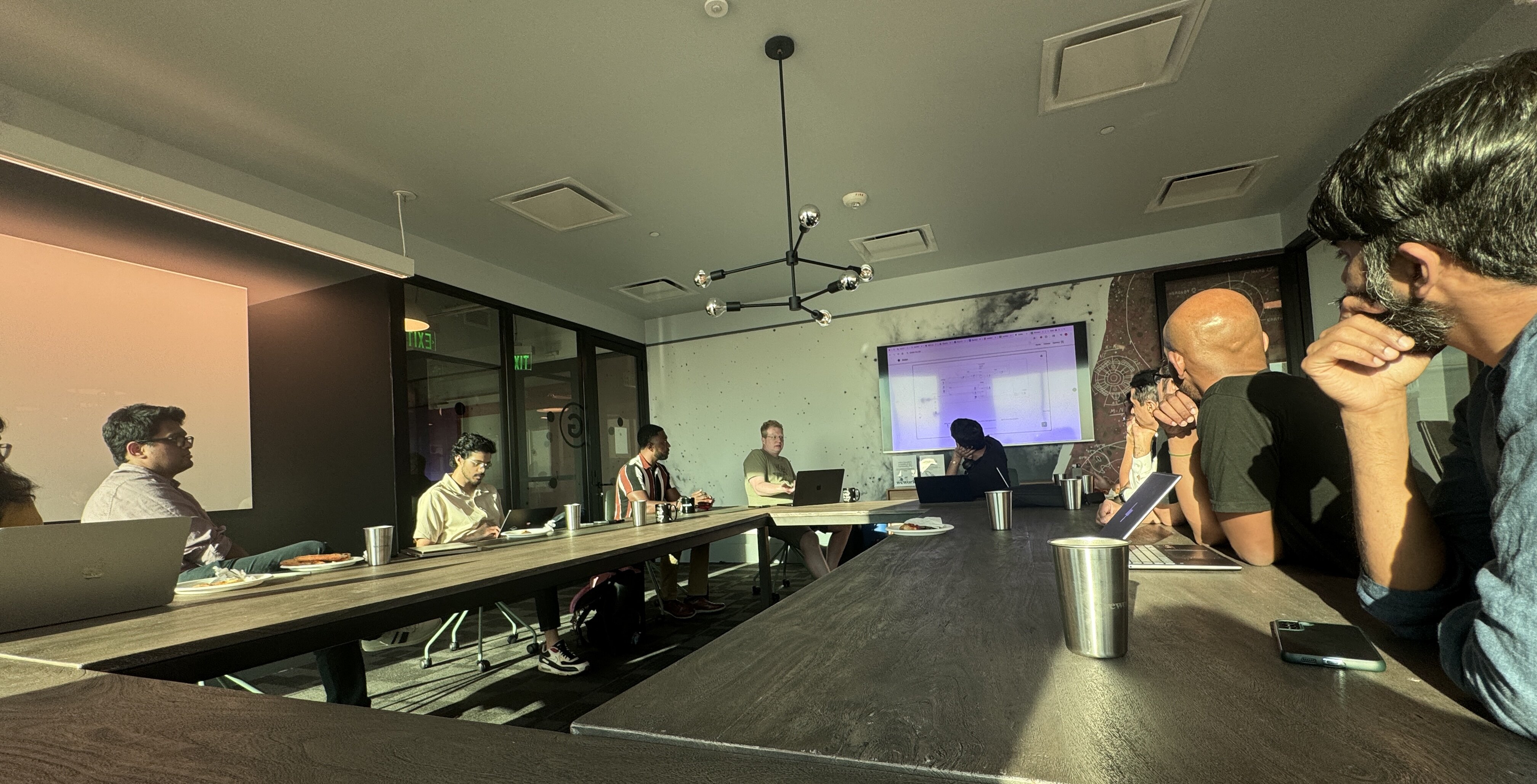 A group of approximately eight people are seated around a large wooden conference table in a modern office meeting room, engaged in a discussion or work session. The room features dramatic, high-contrast lighting, with strong sunlight streaming in from the left, casting long shadows across the table and highlighting the attendees and their open laptops.