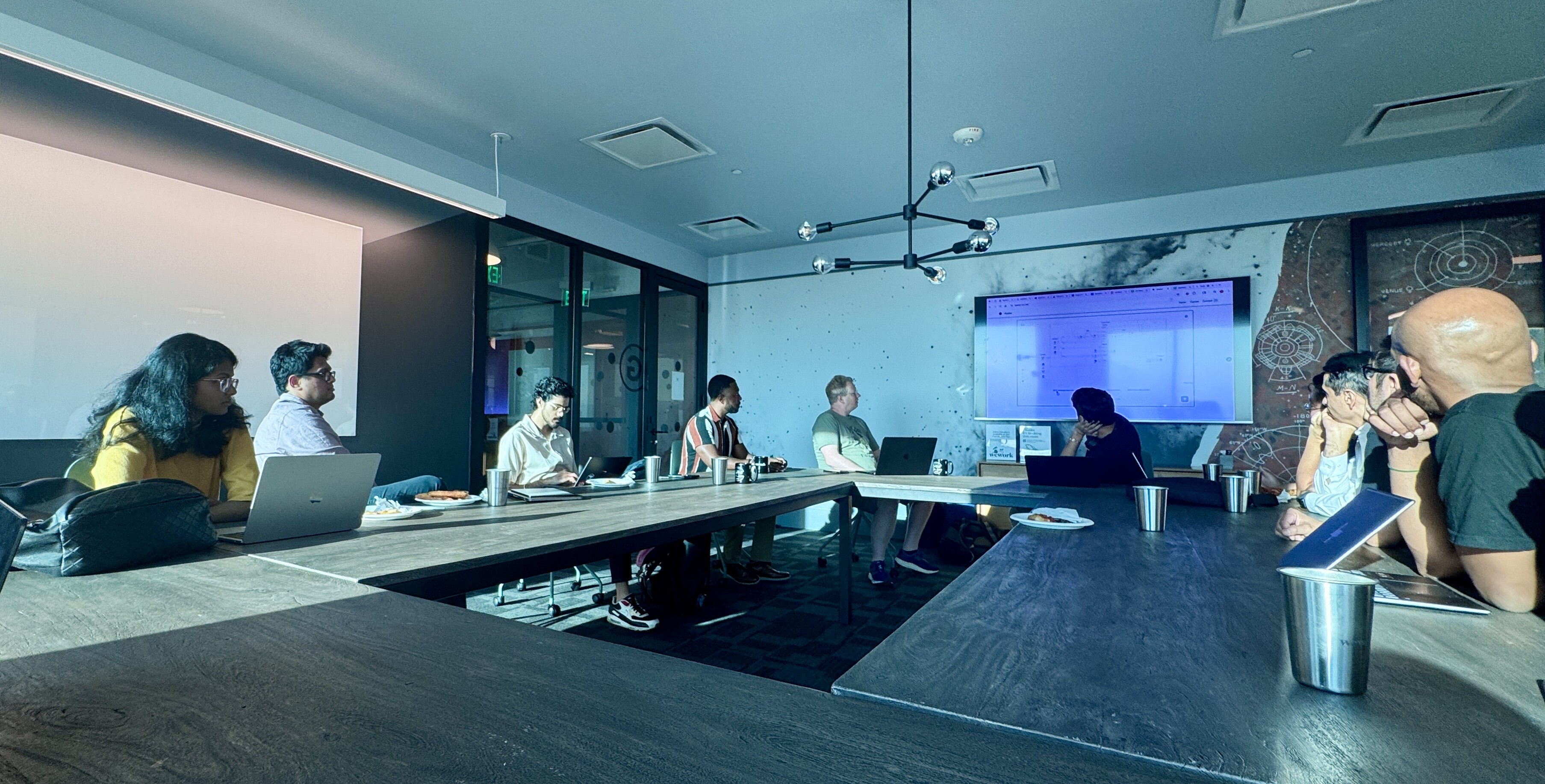 A wide-angle photograph capturing a group of eight people attending a meeting or working session around a large U-shaped conference table in a modern office room. A large screen displaying data or a diagram is visible on the back wall, which is decorated with a dark, map-like mural.