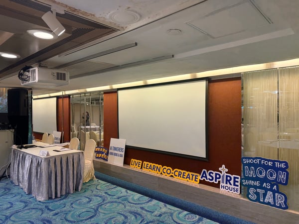 A photograph of an indoor event or seminar room featuring a large projector screen, a draped table, and several motivational signs.