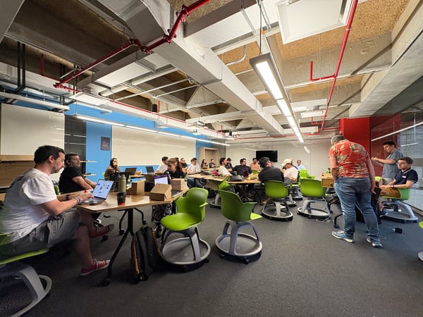 A group of people are gathered in a modern, industrial-style workspace or classroom, many seated at tables with laptops and using bright green ergonomic chairs.
