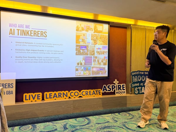 A man stands on a stage holding a microphone, giving a presentation in front of a large screen titled 'AI TINKERERS'.