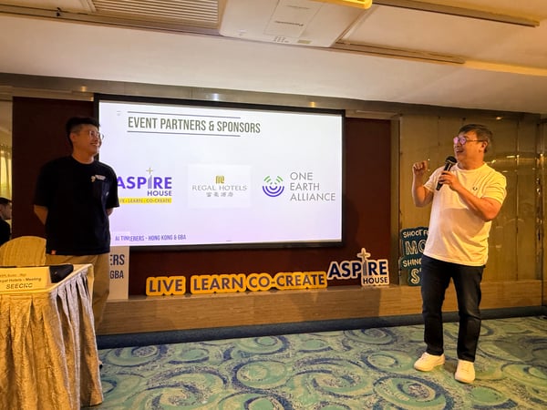 A man speaks into a microphone during a presentation about event partners and sponsors, while another man stands nearby in an indoor setting.