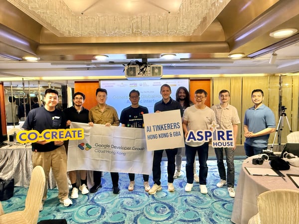 A group of nine men posing for a group photo at an AI hackathon event, holding banners and signs for various tech organizations.