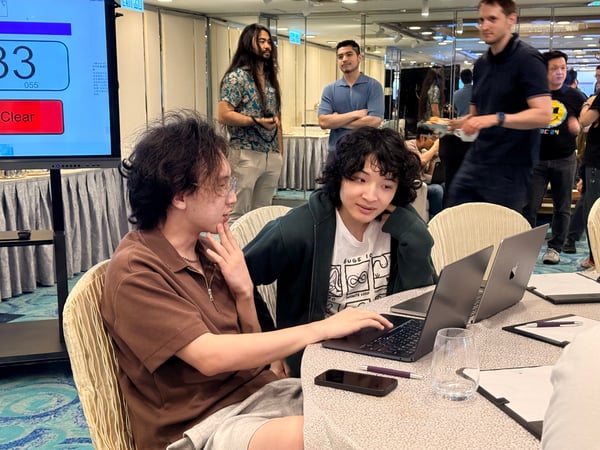 Two individuals are seated at a table with laptops, engaged in a discussion or collaborative work during an indoor event.
