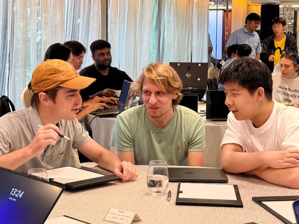 A group of individuals are seated around a table in a well-lit indoor setting, likely a workshop or meeting, engaged in discussion with laptops and tablets.
