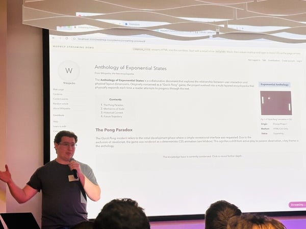 A man is giving a technical presentation in an indoor setting, standing next to a large projection screen that displays a web application demo.