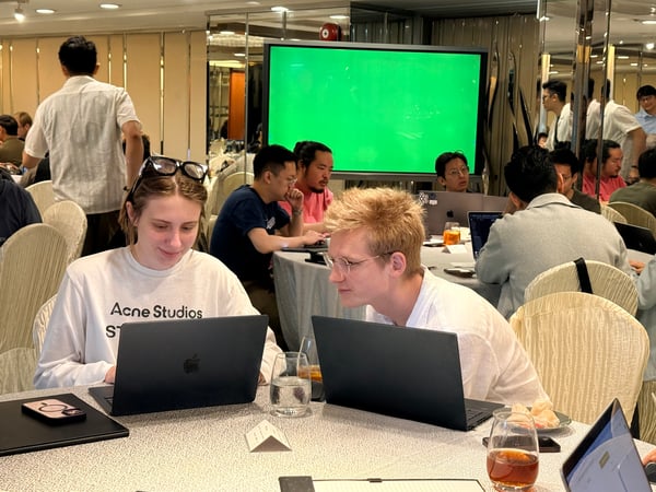 A group of people are seated at round tables in a conference room, with two individuals in the foreground focused on their laptops.