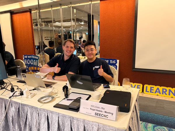 Two men are sitting at a table with laptops, giving a thumbs-up gesture in a meeting room setting.