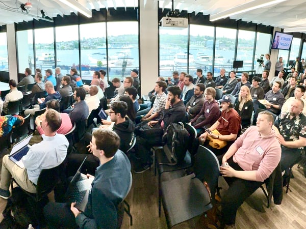 A photograph capturing an indoor technology event where a speaker addresses a large, seated audience in a modern venue overlooking a waterfront cityscape.