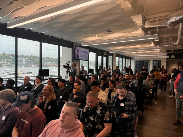 A photo capturing a large audience attending an indoor technology or AI-focused event, seated and standing in a modern space with large windows overlooking a waterfront cityscape.
