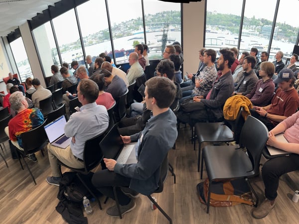 A wide-angle photograph capturing a large audience attending a panel discussion or presentation in a modern event space with large windows overlooking a waterfront city view.