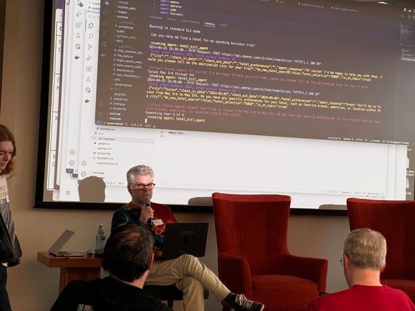 A man is presenting during an indoor technical event, sitting and speaking into a microphone in front of a large projection screen displaying code and conversational API output related to a travel agent bot demo.