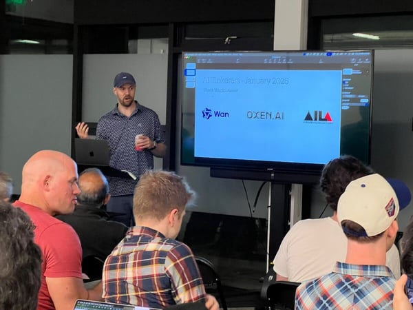 A candid indoor photograph capturing a man giving a presentation to an audience, with the screen displaying a slide titled "AI Tinkerers - January 2026" and several sponsor logos.