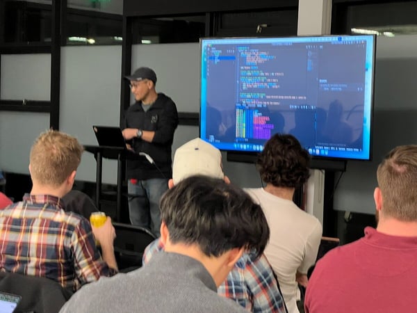 A man wearing a cap is giving a technical presentation, standing next to a lectern while displaying lines of code on a large monitor behind him, to an audience seated in the foreground.