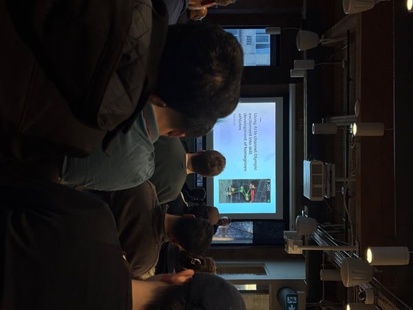 A photograph taken from behind an audience watching a presentation on a large screen in a dimly lit indoor setting.