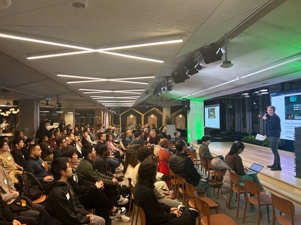 A wide-angle photograph capturing a large audience seated in a modern, dimly lit indoor venue, watching a male presenter on a low stage next to two projection screens displaying presentation slides.
