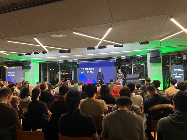 This is a photograph of a large audience attending a tech presentation titled "3D Models with LLMs," featuring two presenters standing on a stage in a modern venue with green accent lighting and large windows overlooking a nighttime city view.