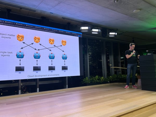 A man wearing a black Spotify t-shirt and jeans is giving a presentation on a wooden stage, standing next to a dark podium, while a large screen displays a technical diagram using emojis of people and robots.