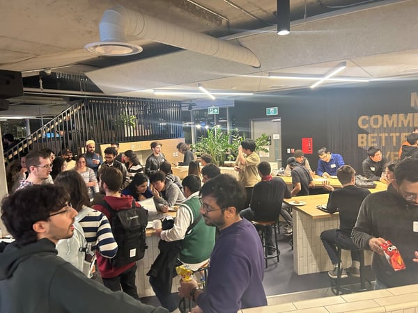 A candid photograph captures a large group of people socializing and seated at high tables in a modern, industrial-style indoor venue, likely during an evening event or gathering.