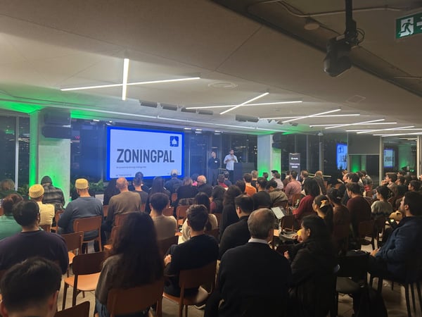 A photograph capturing a large audience seated indoors, attending a presentation where two speakers stand near a stage displaying the company name "ZONINGPAL" on a large screen.