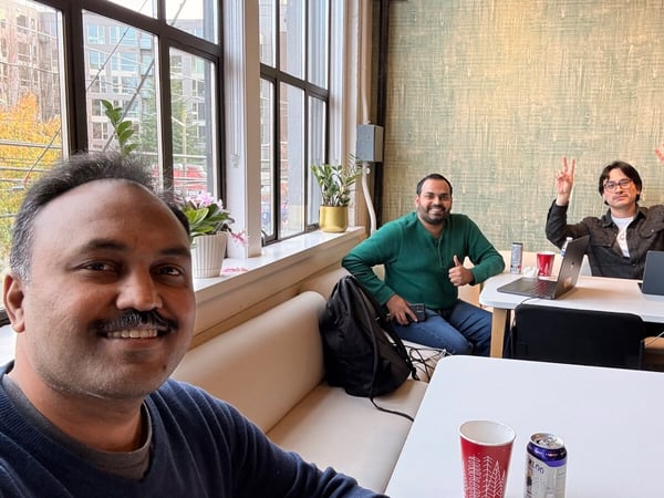 A group selfie taken indoors shows three men smiling and posing at a white table in a modern office space, featuring large industrial-style windows overlooking an urban area.