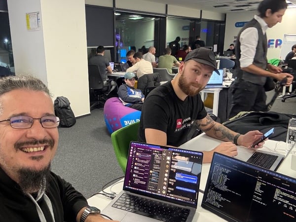 A candid selfie taken in a busy, modern office environment late at night, showing two men smiling while working on open laptops displaying code and chat applications, with many other people working in the background.