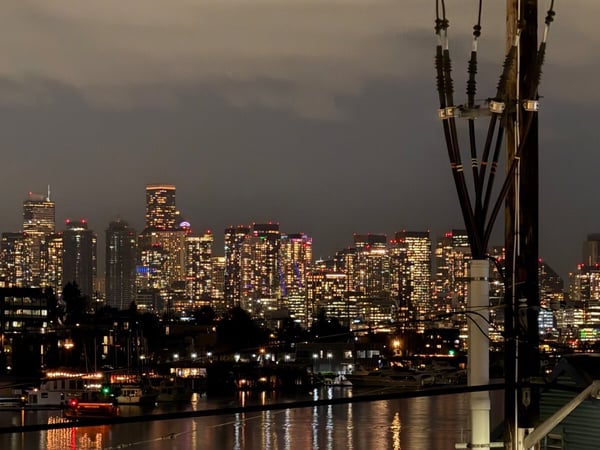 A nighttime photograph captures a dense, illuminated cityscape across a body of water, with numerous high-rise buildings sparkling under a dark, cloudy sky. A utility pole with thick cables stands prominently in the right foreground.