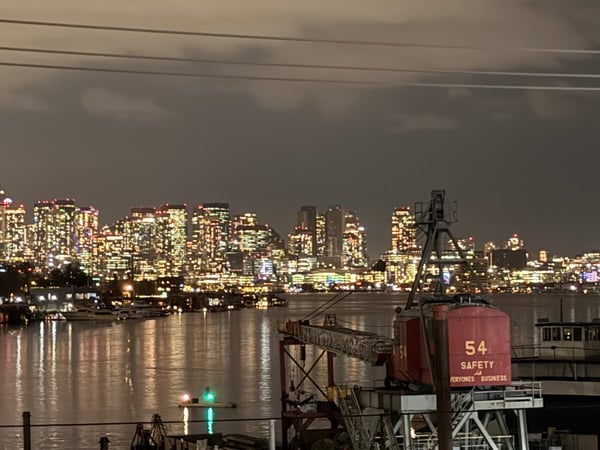 A nighttime photograph captures a brightly lit urban skyline, identifiable as Seattle (featuring the Space Needle), reflecting across a body of water, with a large red industrial crane structure prominent in the foreground.