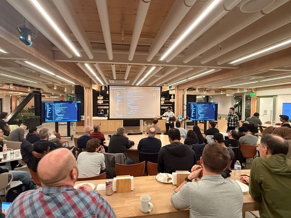 A wide-angle photograph captures a large group of people attending a technical presentation or coding demonstration in a modern, brightly lit indoor event space featuring wooden ceilings and linear lighting.