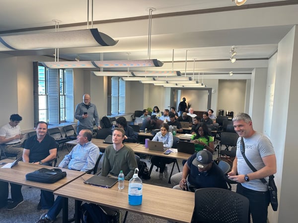 A candid photograph captures a diverse group of approximately 28 people attending an indoor workshop or training session, seated at long wooden tables with many using laptops. Several attendees in the foreground and middle rows are looking toward the camera, including a man smiling broadly on the far right.