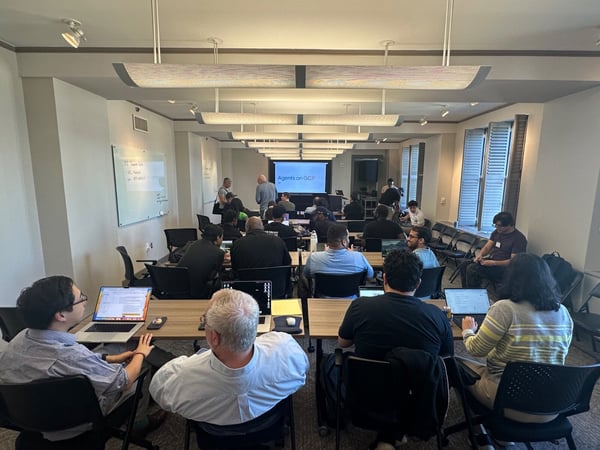 A photograph capturing a technical workshop or class setting where attendees are seated at desks with laptops open, facing a large screen displaying the title "Agents on GCP."
