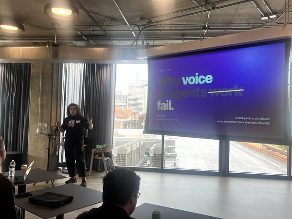 A presenter stands next to a large projection screen displaying a presentation slide about voice AI agents in an indoor event space.