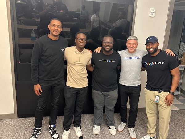 A group photo of five men standing shoulder-to-shoulder and smiling in an indoor office or conference setting, several of whom are wearing branded apparel related to Google and AI groups.